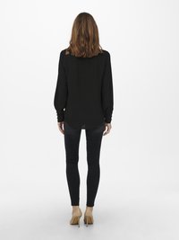 Blusa preta de manga longa com um corte descontraído, tecido texturizado e punhos abotoados, combinada com jeans skinny escuros e sapatilhas nude de salto alto.