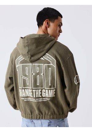 Homme portant un sweat à capuche vert olive avec un grand texte "1980 Change The Game" dans le dos et un jean bleu clair sur fond uni.