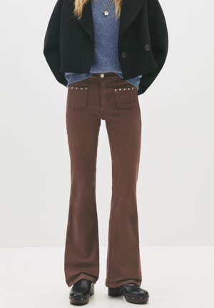 Kobieta nosząca brązowe rozszerzane spodnie z ćwiekowanymi przednimi kieszeniami, czarną krótką kurtkę, niebieski sweter, czarne chodaki i warstwowe naszyjniki.