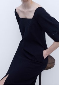 Robe noire ample avec un décolleté carré, des manches trois-quarts et une fente latérale ; dotée d'un tissu doux et texturé ainsi que de poches cachées.