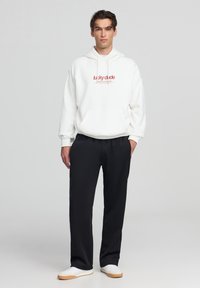 Felpa bianca in cotone con logo rosso al centro, abbinata a pantaloni neri larghi e sneakers bianche con suole chiare.