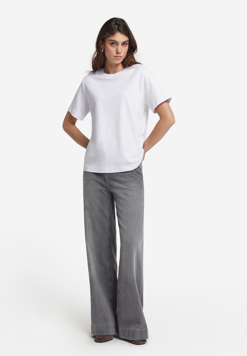 Frau mit braunen Haaren, die ein schlichtes weißes Kurzarmshirt und weit geschnittene graue Jeans trägt, mit Händen an den Hüften.