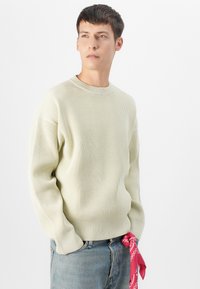 Pulover ribbed de culoare verde deschis cu guler rotund, având mâneci lungi. O bandană roșie legată în talie adaugă un accent contrastant.
