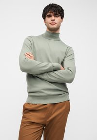 Πουλόβερ ζιβάγκο μέντας σε απαλό υλικό, με εφαρμοστή γραμμή, ριμπ μανσέτες και αδιάκοπος γιακάς σε ρολό. Ορατό καφέ παντελόνι.