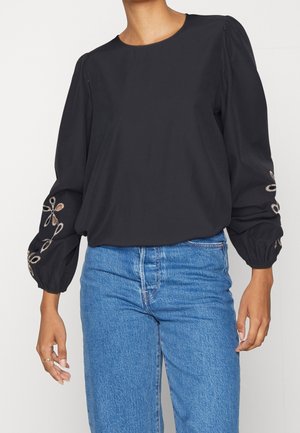 Woman wearing a black long-sleeve blouse with puffed sleeves and embroidered floral details paired with blue jeans.