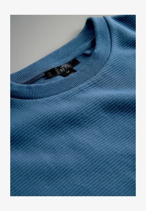 Sinine soonikkoes pusa, millel on ümara kaelusega ja sees kaelas nähtav must silt tekstiga "THE SET IM".