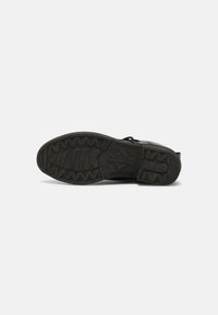 Semelle de chaussure noire avec des motifs de sculptures, incluant des formes en zigzag et en losange, présentée à plat sur un fond uni gris clair.