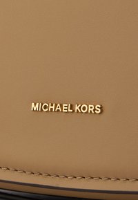 Materiale sintetico marrone chiaro con una texture liscia e logo dorato "MICHAEL KORS" in rilievo. Design semplice senza ulteriori accessori o motivi.