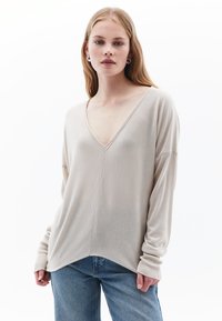 Suéter de manga larga beige con cuello en V, de tejido suave, hombros caídos y puños acanalados. Combinado con pantalones de mezclilla azul claro.