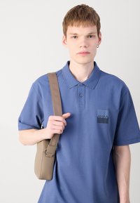 Blå kortärmad polo med logotyp, kombinerad med en beige axelväska. Mjuk tygkänsla, klassisk krage med knappslå.