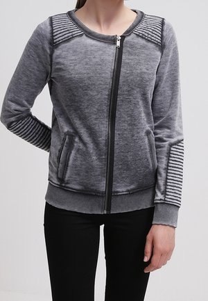 Kvinde iført en grå lynlåstrøje med ribbet polstrede detaljer på skuldre og ærmer, kombineret med sorte bukser.