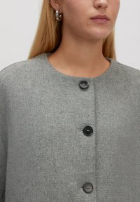 Frau mit langen blonden Haaren trägt einen grauen Pullover mit drei schwarzen Knöpfen und silbernen Creolen, neutraler Hintergrund.