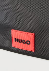 Μαύρο υφασμάτινο υλικό με κόκκινο ορθογώνιο έμπλαστρο που εμφανίζει τη λέξη "HUGO" με ανάγλυφα μαύρα γράμματα.