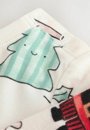 Fleece-tyg med en ljus krämfärgad bakgrund, med ett leende grön-randig träddesign och röda-svart detaljer. Tryckknapp detalj synlig.