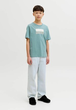 Majica svetlo zelene barve z belim logotipom, kombinirana z lahkimi modrimi kavbojkami s ravnimi hlačnicami in črnimi semišnimi čevlji. Preprost dizajn, sproščen kroj.