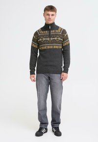 Mørkegrå strikket sweater med høj krave, der har et geometrisk mønster i gul og tan. Båret sammen med grå jeans og sorte sneakers.