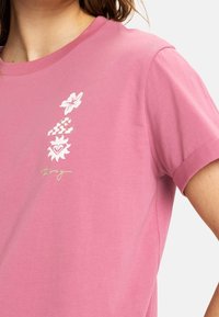 Růžové bavlněné tričko s květinovými a srdíčkovými potisky v bílé barvě a vlnkovým designem. Krátké rukávy, kulatý výstřih a pohodlný střih.