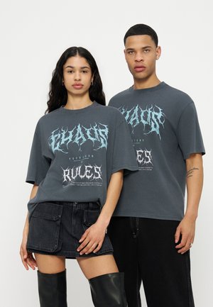 Mladý muž a žena stojaci, obaja majú na sebe tmavé tričká s potlačou; žena má čiernu sukňu a čižmy nad kolená, muž čierne nohavice.