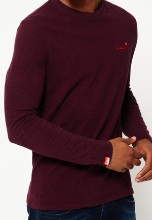 Man draagt een nauwsluitend bordeauxrood shirt met lange mouwen met een klein rood logo op de borst en pols, gecombineerd met donkerblauwe spijkerbroek.