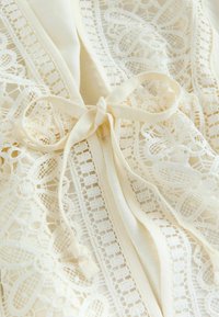 Haină de culoarea cremei cu detalii delicate din dantelă, având închidere cu panglică și modele florale intricate, evidențiind textura și designul.