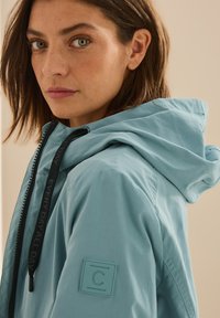 Veste imperméable bleu clair avec capuche, dotée d'un tissu texturé, de cordons de serrage noirs et d'un écusson logo sur la manche.