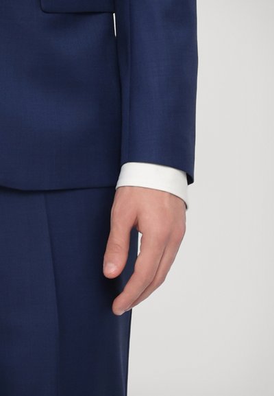 Gros plan sur une veste de costume bleu marine avec un tissu lisse, un poignet de chemise blanche qui dépasse, et une main détendue placée sur le côté.