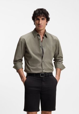 Hombre con cabello rizado y oscuro, vistiendo una camisa verde oliva abotonada con las mangas remangadas y pantalones cortos negros con cinturón, con las manos en los bolsillos.