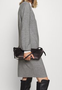 Robe grise à manches longues et coupe décontractée, associée à une pochette en cuir marron foncé, avec une sangle nouée et une texture lisse.