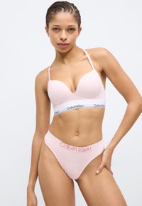 Ανοιχτό ροζ bralette Calvin Klein με λεπτές τιράντες και λαιμόκοψη σε σχήμα V, συνδυασμένο με ταιριαστά σλιπ με λογότυπο στη μέση. Υφασμάτινη επιφάνεια με ομαλή υφή.