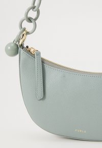 Sac à main en cuir bleu clair avec une forme incurvée, fermeture éclair dorée et une bandoulière en chaîne. Présente un accent rond vert près de la fermeture éclair.