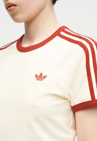 Gros plan d'une personne portant un t-shirt crème avec des bordures rouges et trois bandes rouges sur l'épaule, arborant un petit logo rouge sur la poitrine.