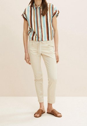 Mujer que lleva una camisa de manga corta abotonada con rayas verticales en azul, marrón y blanco, pantalones beige con cordón y sandalias marrones abiertas en los dedos.