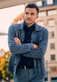 Gestructureerde blauwgrijze geweven jas met een kraag en knoopdetails, gedragen over een donker overhemd, gecombineerd met grijze broek.