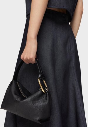 Femme tenant un sac à main en cuir noir texturé avec des ferrures dorées, portant une jupe en denim bleu foncé et un haut court sur fond gris clair.