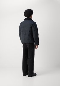 Sort pufferjakke med vandret quiltning og sort krave, båret over mørke bukser og sorte sko. Ensfarvet baggrund.