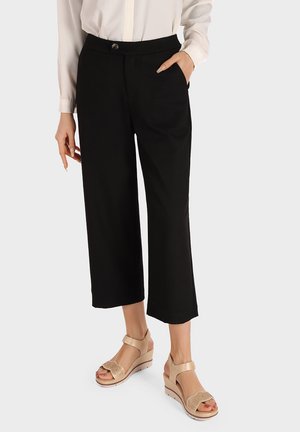 Pantaloni neri cropped con tasche laterali, texture liscia e un bottone in vita, abbinati a sandali con piattaforma beige caratterizzati da accenti floreali.