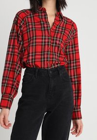 Chemise à carreaux rouge et noir à boutons, manches longues, avec col et manches retroussées, portée avec un jean noir taille haute.