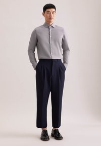 Maglietta grigia a maniche lunghe con chiusura a bottoni, abbinata a pantaloni pieghettati blu navy. Il modello indossa scarpe nere con lacci. Sfondo neutro.