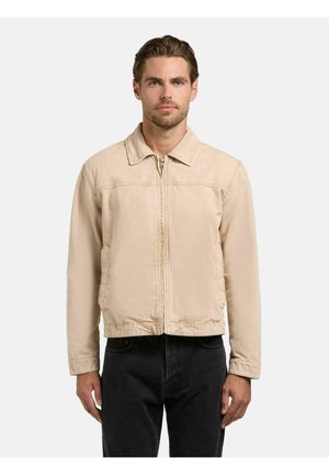 Mann med kort brunt hår iført en beige genser med glidelås og svarte bukser, stående foran en ensfarget lys bakgrunn.