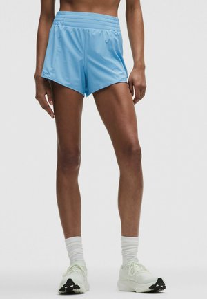 Persona atlética con pantalones cortos de correr azul claro, calcetines blancos y zapatillas blancas, de pie frente a un fondo liso.