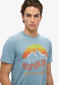 Camiseta azul jaspeada con un estampado gráfico de montañas y un atardecer. El texto dice "SUPERDRY" en negrita roja y amarilla, con un corte relajado.