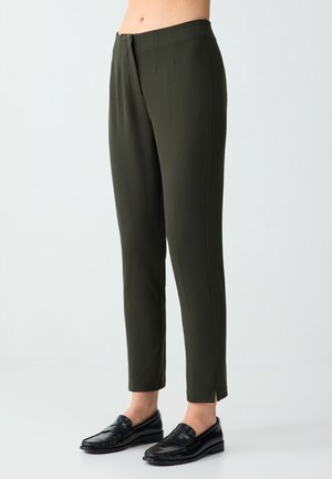 Ciemnozielone spodnie slim fit z przednimi zakładkami i małymi bocznymi rozcięciami, noszone z czarnymi skórzanymi mokasynami na jasnym tle.