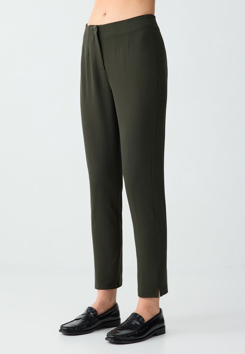 Pantalon slim vert foncé avec plis devant et petites fentes latérales, porté avec des mocassins en cuir noir sur fond clair.