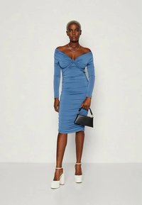 Vestido azul sem ombros com mangas longas e detalhe de franzido, até à meia panturrilha. Acompanhado de uma pequena mala preta e sapatos de salto branco.
