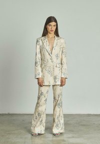 Completo beige con motivo floreale, composto da un blazer su misura e pantaloni a zampa d'elefante, con accenti floreali blu e verdi, realizzato in tessuto liscio.