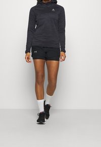 Felpa nera a maniche lunghe con tessuto testurizzato e cappuccio, abbinata a pantaloncini sportivi neri. Calzini bianchi alla caviglia e scarpe sportive nere.