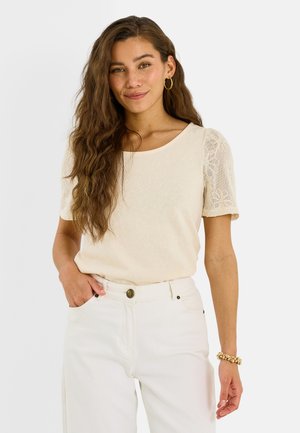 Femme aux longs cheveux ondulés portant un haut beige à manches en dentelle, un pantalon blanc, des boucles d'oreilles créoles dorées et un bracelet en or, debout avec une main dans la poche.