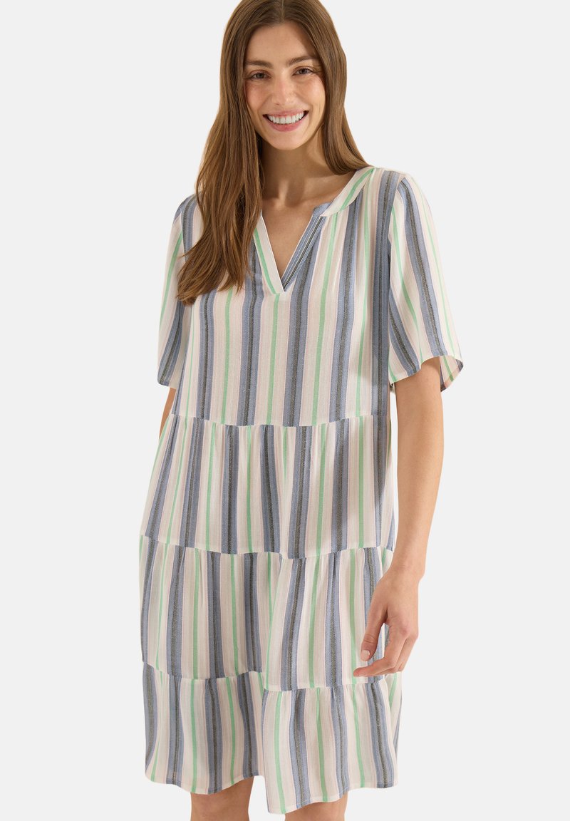 Femme souriante portant une robe ample à manches courtes, longueur genou, avec des rayures verticales bleues, vertes et blanches sur un fond clair.