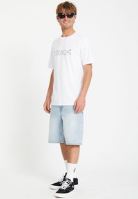T-shirt blanche avec logo "Volcom" noir, shorts en denim bleu clair, baskets noires avec accents blancs et lunettes de soleil noires.