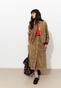 Veste en velours côtelé marron et jupe assortie. Haut rouge en dessous. Boutons sur le devant de la jupe. Mocassins noirs et chaussettes grises. Sac à carreaux tenu sur le côté.